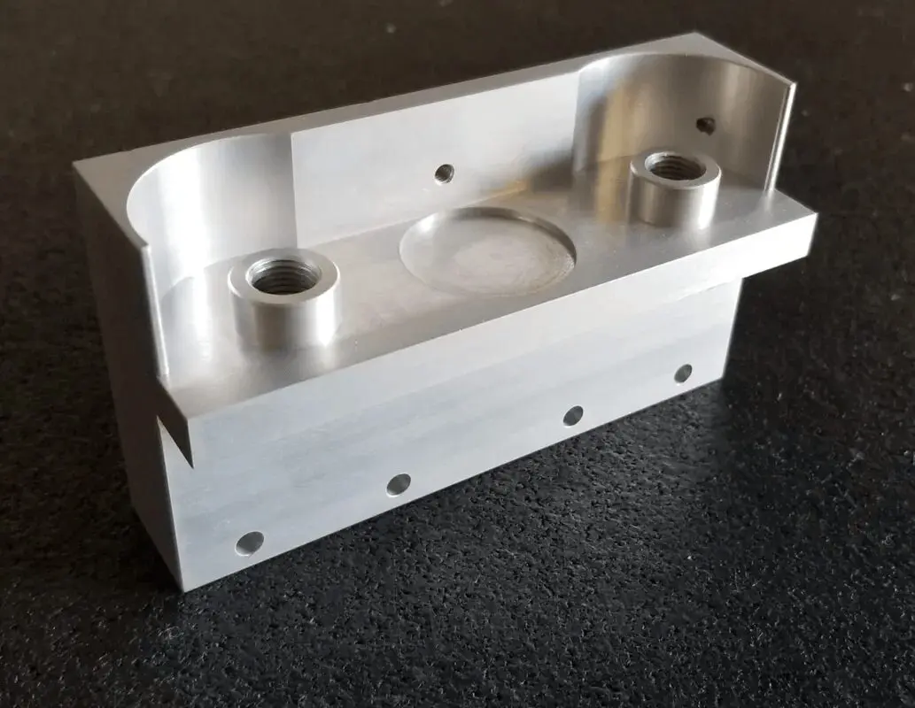 Metallic rectangular component with a central recessed circular area, two threaded holes, and multiple smaller holes along the bottom edge, set against a dark background.